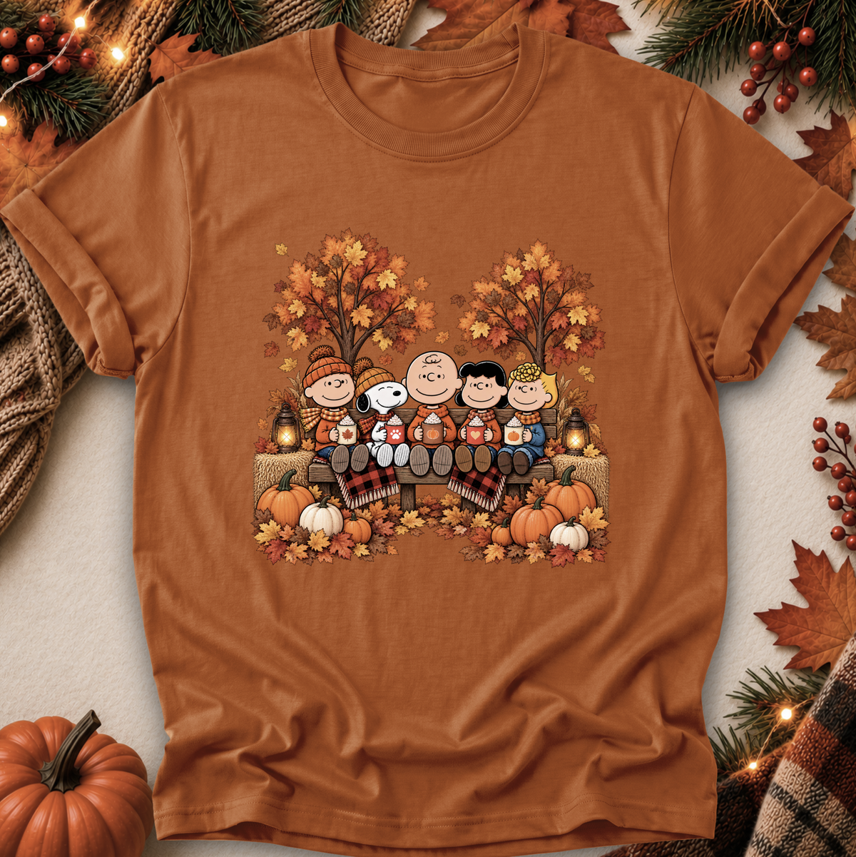 A rust-orange crewneck t-shirt featuring a colorful graphic of the Peanuts characters sitting by a campfire during autumn. The shirt is styled in a flat-lay with seasonal props including pumpkins, pine branches, and fall leaves.