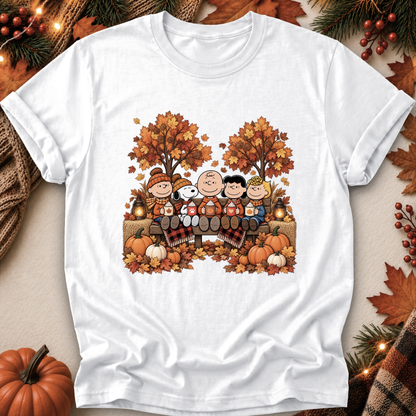 A white short-sleeve crewneck t-shirt featuring a colorful autumn-themed graphic of Peanuts characters sitting in a wagon with pumpkins and fall trees. The shirt is displayed flat on a cozy, styled background with autumn leaves, pine branches, and a small pumpkin.