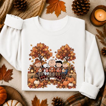 A white long-sleeve sweatshirt laid flat on a cozy autumn-themed background. The front of the sweatshirt features a colorful cartoon graphic of Peanuts characters sitting on a bench surrounded by fall trees, pumpkins, and leaves.