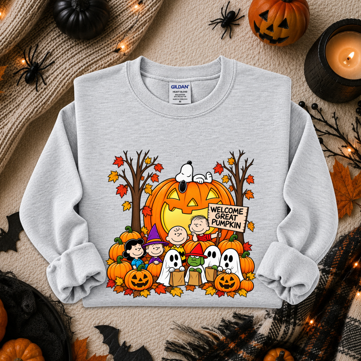 A light gray long-sleeve sweatshirt with a colorful 'Welcome Great Pumpkin' Peanuts graphic, styled in a flat lay on a beige knit blanket with Halloween props including pumpkins, a candle, and spider decorations.