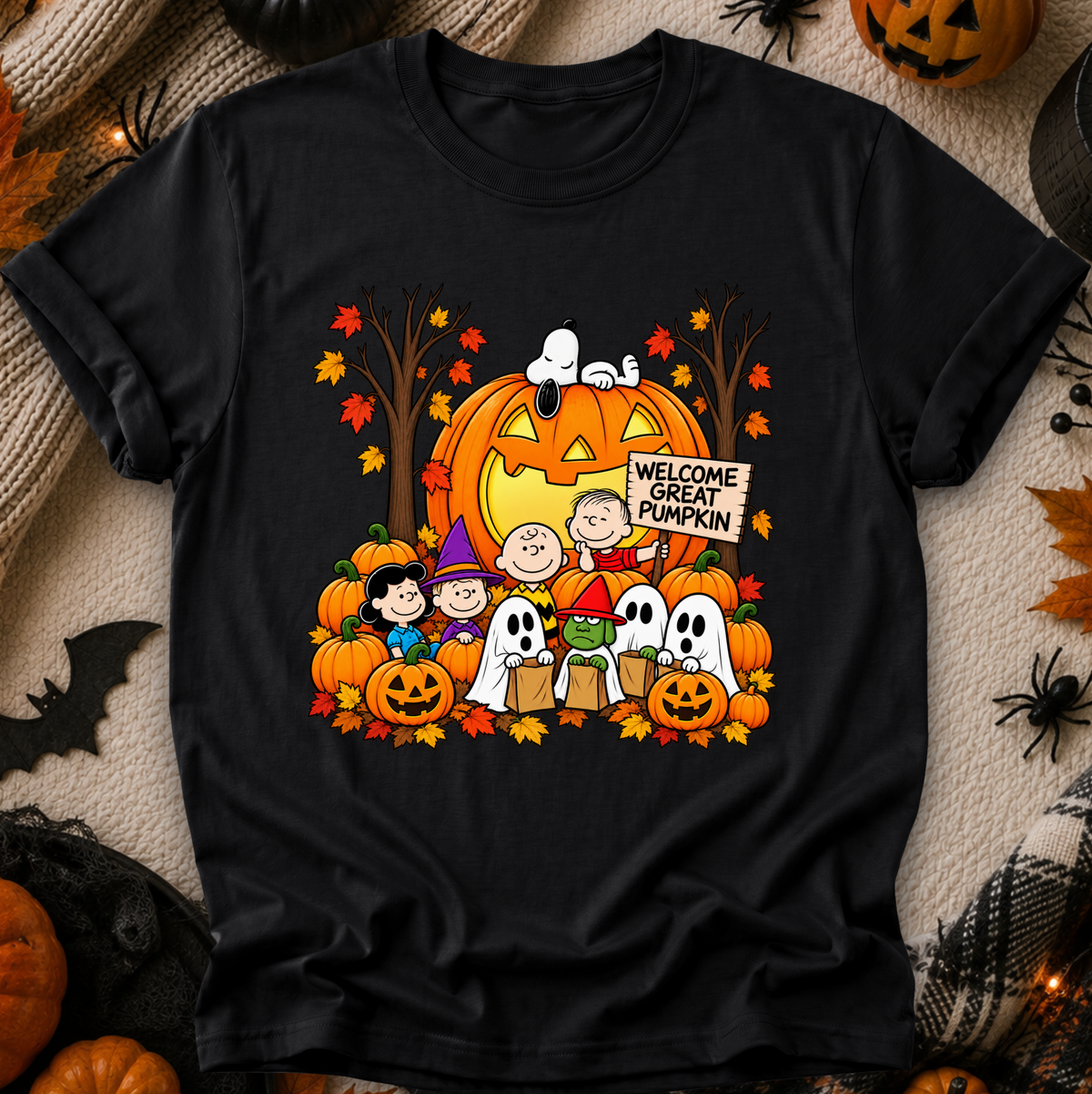 A black short-sleeve graphic T-shirt laid flat on a cozy beige fabric background with Halloween props. The shirt features a colorful cartoon print of Snoopy, Woodstock, Charlie Brown, and the Peanuts characters with pumpkins, ghosts, autumn leaves, and a sign reading 'Welcome Great Pumpkin.