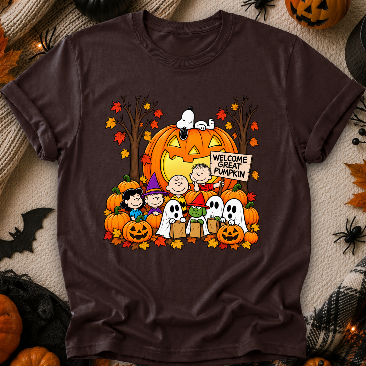 A dark brown short-sleeve crewneck t-shirt featuring a colorful graphic of Peanuts characters in Halloween costumes in front of a jack-o'-lantern with a 'Welcome Great Pumpkin' sign. The shirt is displayed flat on a cozy beige knit blanket surrounded by Halloween props like pumpkins, a spider, and a bat.