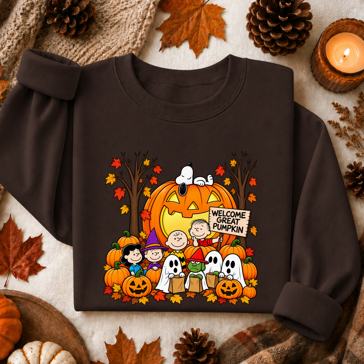 A dark brown long-sleeve sweatshirt featuring a colorful cartoon graphic of the Peanuts characters in a Halloween scene with pumpkins, ghosts, and a sign reading 'Welcome Great Pumpkin.' The sweatshirt is styled in a flat lay with autumn leaves, pinecones, a candle, and knitted fabric around it.