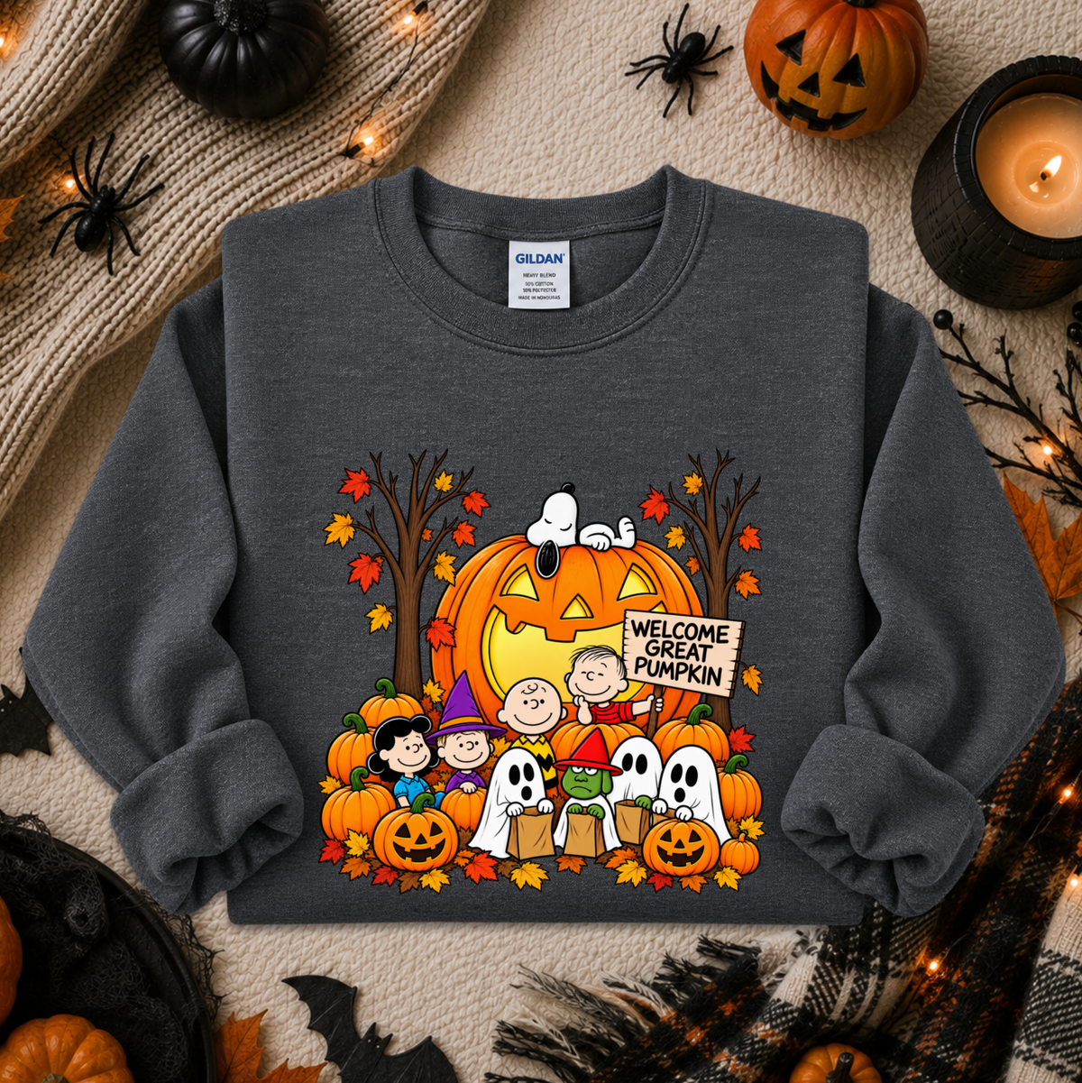 A dark gray crewneck sweatshirt featuring a colorful 'Welcome Great Pumpkin' Peanuts graphic, styled in a cozy Halloween flat-lay with pumpkins, a candle, and autumn decorations.