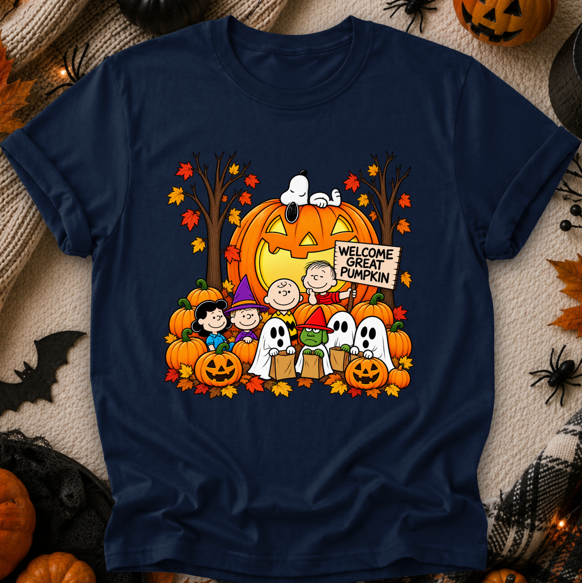 A navy blue crewneck t-shirt featuring a colorful cartoon graphic of the Peanuts characters with pumpkins, ghosts, and a jack-o'-lantern holding a 'Welcome Great Pumpkin' sign, displayed on a cozy autumn-themed background.