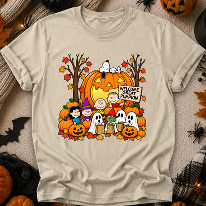A beige crewneck t-shirt featuring a colorful graphic of the Peanuts characters Snoopy, Woodstock, Charlie Brown, and Lucy in Halloween costumes, set against a background of pumpkins and autumn leaves. The graphic includes a jack-o'-lantern with a 'Welcome Great Pumpkin' sign.