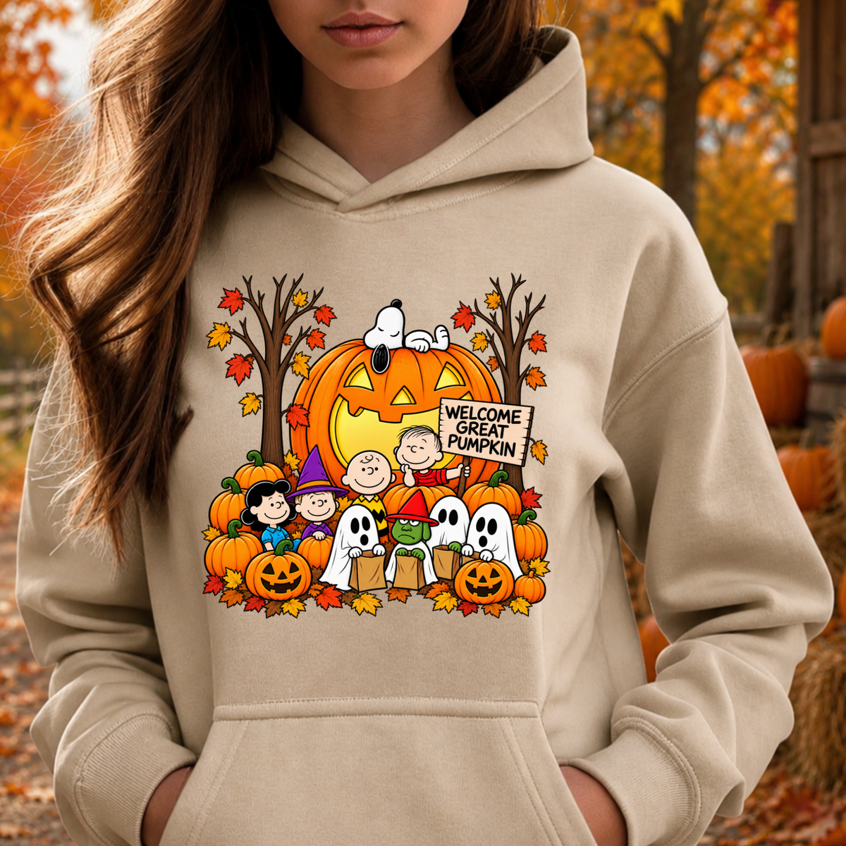 A woman wearing a beige hoodie with a colorful cartoon graphic of Peanuts characters around a jack-o'-lantern pumpkin, photographed outdoors in an autumn setting.