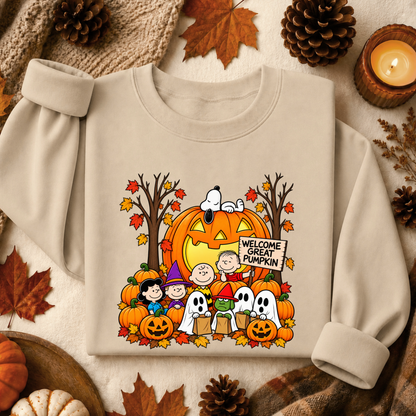A beige crewneck sweatshirt featuring a colorful graphic print of Snoopy and Charlie Brown with pumpkins, ghosts, and a 'Welcome Great Pumpkin' sign, styled in a flat lay with autumn leaves, pinecones, and a candle.