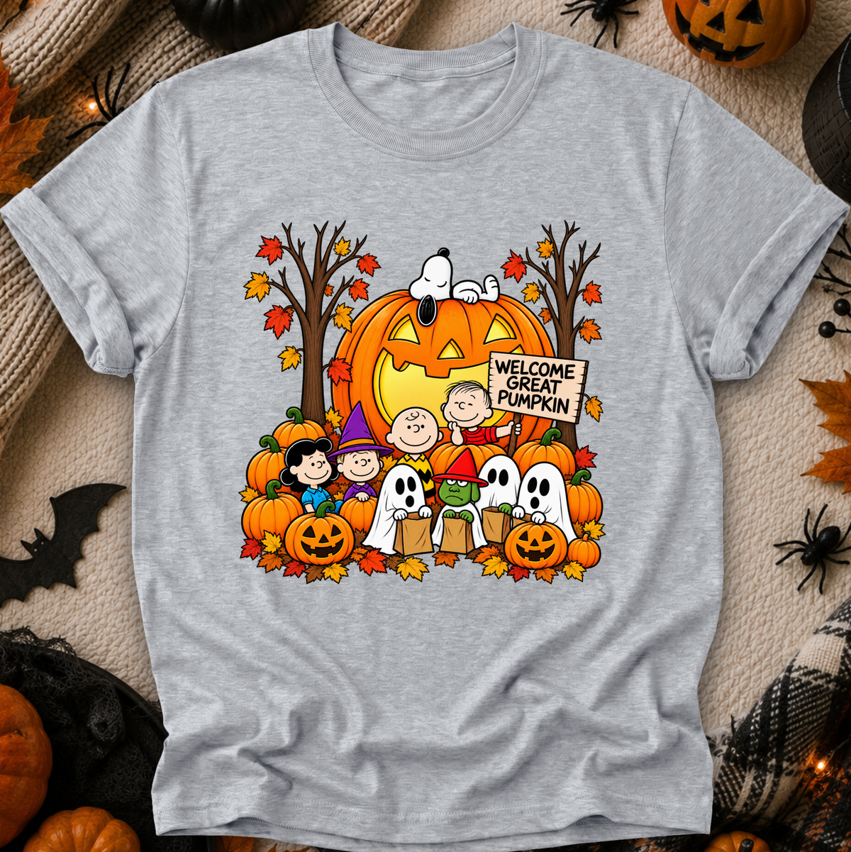 A heather gray short-sleeve T-shirt laid flat with a large colorful Halloween graphic on the front. The print shows Snoopy and Charlie Brown with pumpkins, ghosts, autumn trees, and a sign reading “Welcome Great Pumpkin.