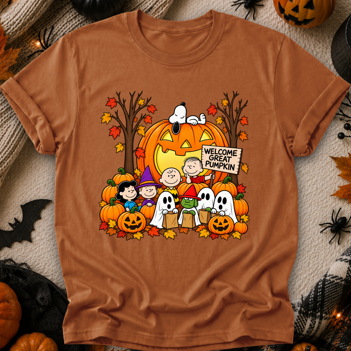 A rust-orange crewneck t-shirt featuring a colorful graphic of Snoopy, Woodstock, Charlie Brown, Lucy, and Snoopy with a jack-o'-lantern and ghosts, surrounded by autumn leaves and pumpkins. The shirt is displayed flat on a cozy, styled background with Halloween decorations.