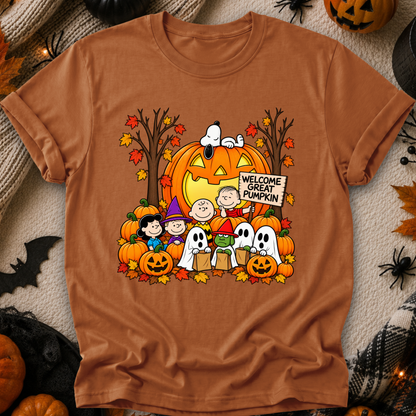 A rust-orange crewneck t-shirt featuring a colorful graphic of Snoopy, Woodstock, Charlie Brown, Lucy, and Snoopy with a jack-o'-lantern and ghosts, surrounded by autumn leaves and pumpkins. The shirt is displayed flat on a cozy, styled background with Halloween decorations.