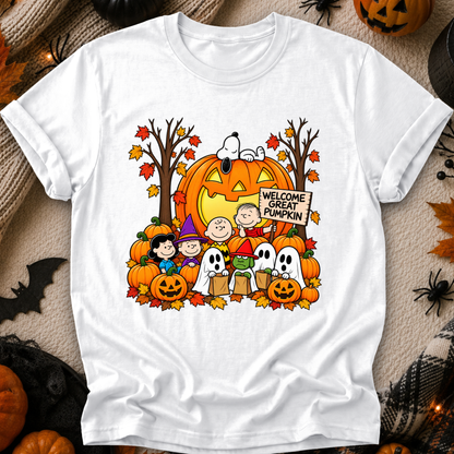 A white short-sleeve crewneck t-shirt featuring a colorful graphic of the Peanuts characters Snoopy, Woodstock, Charlie Brown, and Lucy in an autumn Halloween scene with pumpkins and a 'Welcome Great Pumpkin' sign. The shirt is displayed flat on a cozy, rustic background decorated with pumpkins, spiderwebs, and autumn leaves.