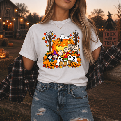 A woman wearing a white graphic t-shirt featuring the Peanuts characters Snoopy, Woodstock, Charlie Brown, and others in a Halloween pumpkin scene with the text 'WELCOME GREAT PUMPKIN'. She has a black and white plaid flannel shirt draped off her shoulders and is wearing blue jeans, set against a warm, rustic autumn outdoor background with pumpkins and a house.