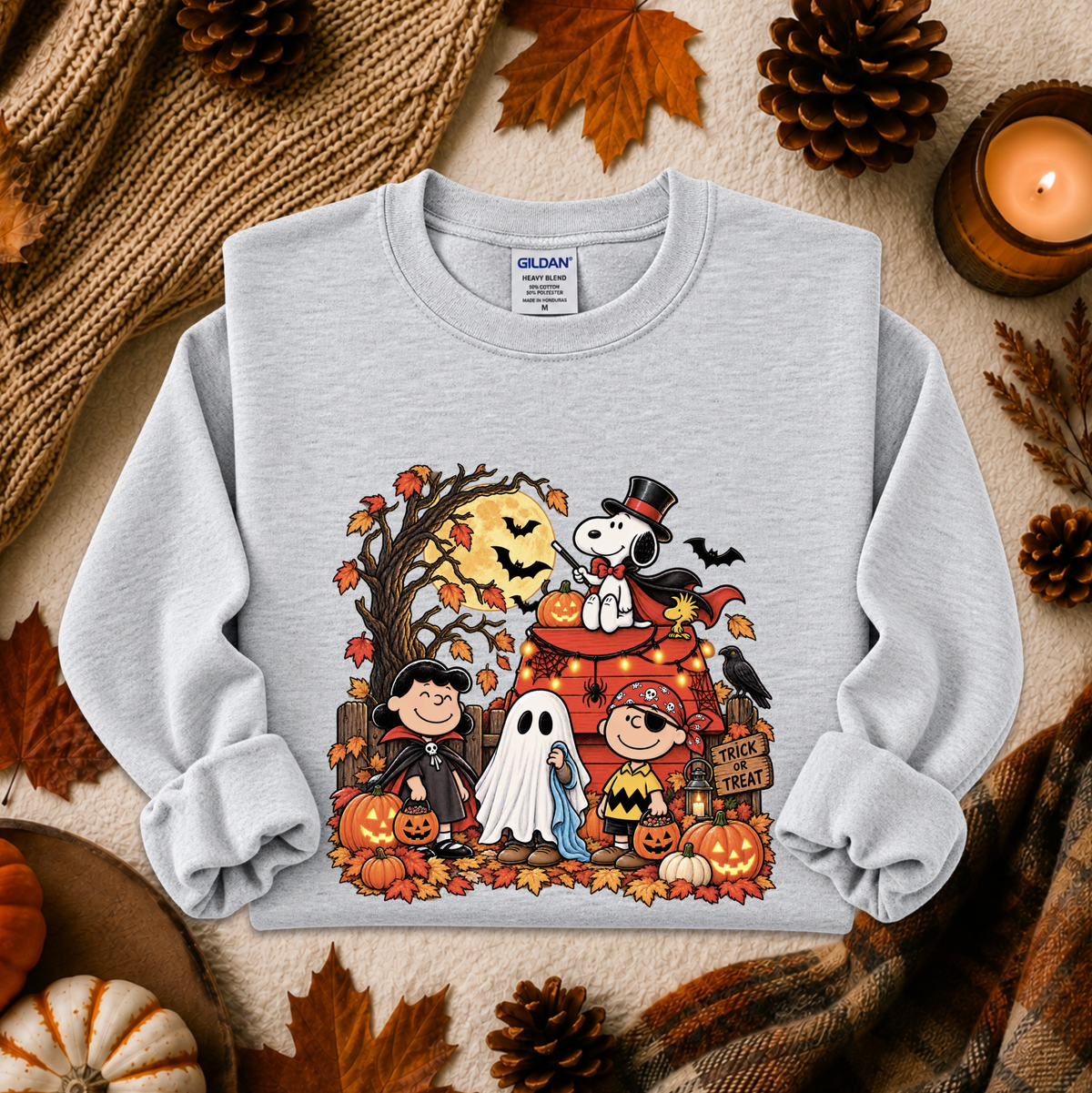 A light gray crewneck sweatshirt featuring a colorful cartoon graphic of Peanuts characters dressed in Halloween costumes, surrounded by pumpkins, ghosts, and autumn leaves. The sweatshirt is styled in a cozy flat-lay setting with a knitted blanket, pinecones, a lit candle, and fall leaves.