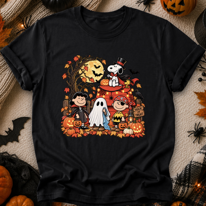 A black short-sleeve crewneck t-shirt featuring a colorful graphic print of Peanuts characters in Halloween costumes, styled flat on a cozy beige knit blanket with autumn props like pumpkins, a spider, and a bat.
