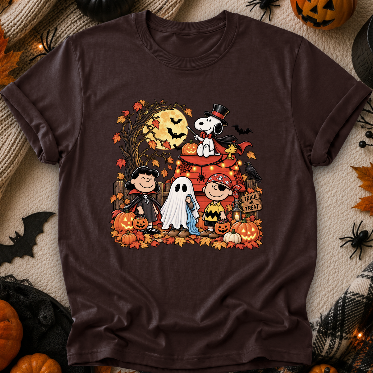 A dark brown short-sleeve crewneck t-shirt featuring a colorful graphic of Peanuts characters in Halloween costumes, styled in a flat-lay with autumn props like pumpkins, a knitted blanket, and black spider decorations.