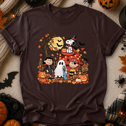 A dark brown short-sleeve crewneck t-shirt featuring a colorful graphic of Peanuts characters in Halloween costumes, styled in a flat-lay with autumn props like pumpkins, a knitted blanket, and black spider decorations.