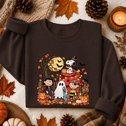 A dark brown long-sleeve sweatshirt laid flat on a neutral fabric background, surrounded by autumn props including pinecones, leaves, a candle, and small pumpkins. The sweatshirt features a colorful Halloween graphic with cartoon characters in costumes, pumpkins, bats, and a ghost.