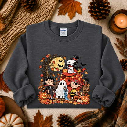 A dark gray crewneck sweatshirt featuring a colorful Halloween graphic of Snoopy, Charlie Brown, Woodstock, Peanuts, a ghost, and pumpkins, styled in an autumn flat-lay with a candle, scarf, and pinecones.