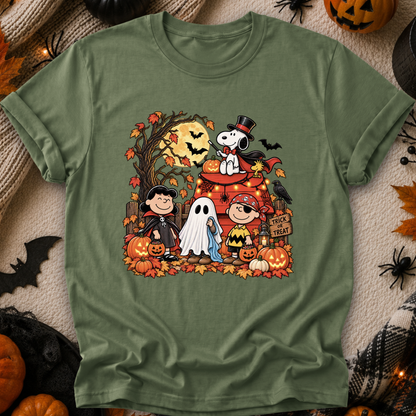 An olive green short-sleeve t-shirt featuring a colorful cartoon graphic of Peanuts characters in Halloween costumes, styled in a flat-lay with autumn and Halloween props.