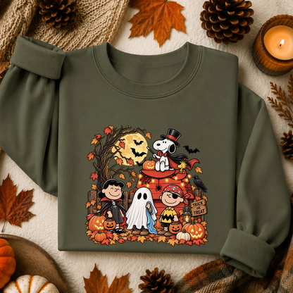 An olive green long-sleeve sweatshirt with a colorful cartoon graphic of Peanuts characters in Halloween costumes, styled in a flat lay with autumn leaves, pinecones, a candle, and pumpkins.