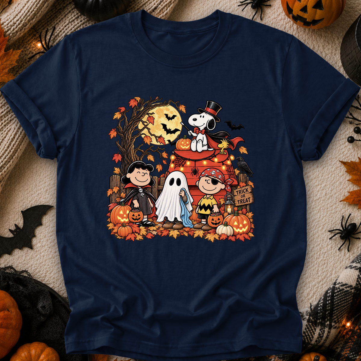 A navy blue crewneck t-shirt featuring a colorful graphic print of Peanuts characters in Halloween costumes, styled flat on a cozy beige knit blanket with autumn props like pumpkins, a spider, and a spiderweb.