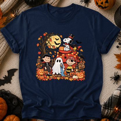 A navy blue crewneck t-shirt featuring a colorful graphic print of Peanuts characters in Halloween costumes, styled flat on a cozy beige knit blanket with autumn props like pumpkins, a spider, and a spiderweb.