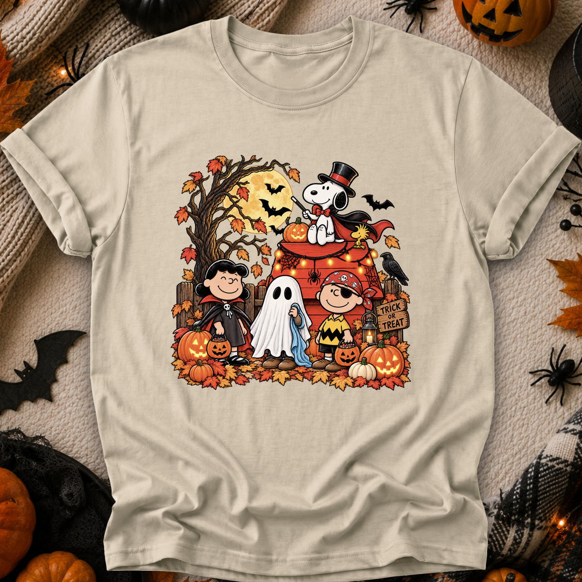 A beige graphic t-shirt featuring a colorful Halloween-themed illustration of Peanuts characters, Snoopy, Woodstock, Charlie Brown, and Lucy, surrounded by pumpkins, bats, and autumn leaves. The shirt is displayed flat on a cozy, themed background with blankets, spider decorations, and jack-o'-lanterns.