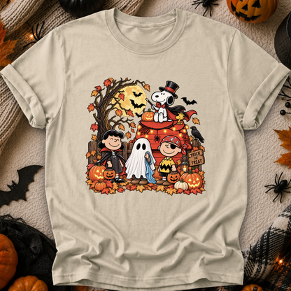 A beige graphic t-shirt featuring a colorful Halloween-themed illustration of Peanuts characters, Snoopy, Woodstock, Charlie Brown, and Lucy, surrounded by pumpkins, bats, and autumn leaves. The shirt is displayed flat on a cozy, themed background with blankets, spider decorations, and jack-o'-lanterns.