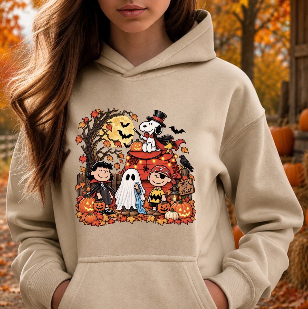 A woman wearing a beige hoodie with a colorful cartoon Halloween graphic featuring Snoopy, Woodstock, Charlie Brown, Peanuts characters, pumpkins, ghosts, and autumn leaves, photographed outdoors in an autumn setting.