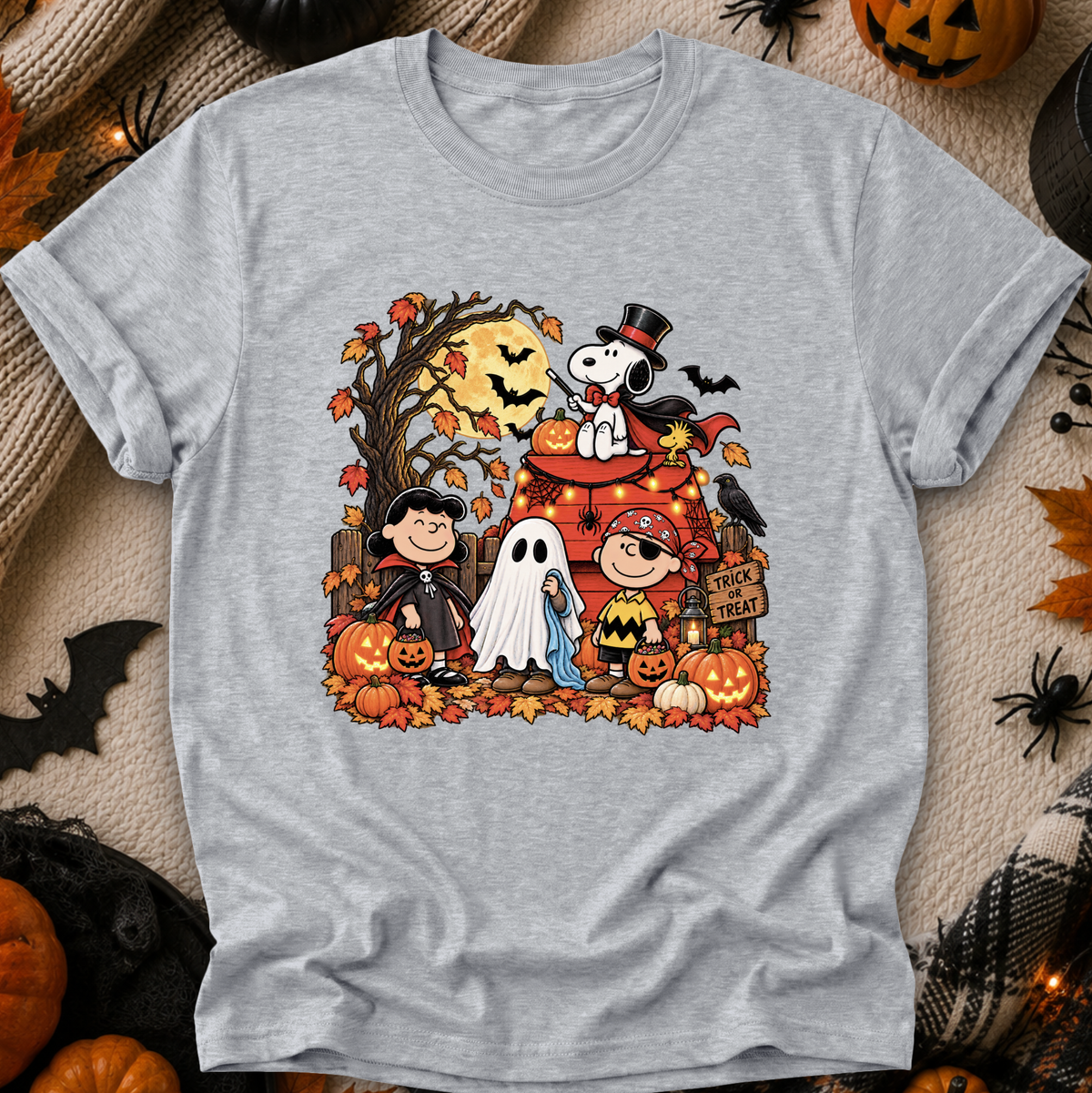 A heather gray short-sleeve t-shirt featuring a colorful graphic print of Peanuts characters in a Halloween scene, laid flat on a cozy beige knit blanket surrounded by autumn props like pumpkins, spider decorations, and a black spiderweb.