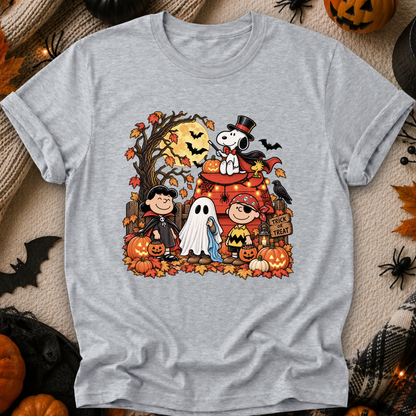 A heather gray short-sleeve t-shirt featuring a colorful graphic print of Peanuts characters in a Halloween scene, laid flat on a cozy beige knit blanket surrounded by autumn props like pumpkins, spider decorations, and a black spiderweb.