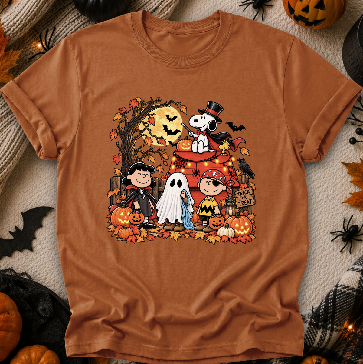 A rust-orange crewneck t-shirt featuring a colorful graphic of Peanuts characters in Halloween costumes, styled in a flat-lay with autumn props like pumpkins, a plaid blanket, and black spider decorations.