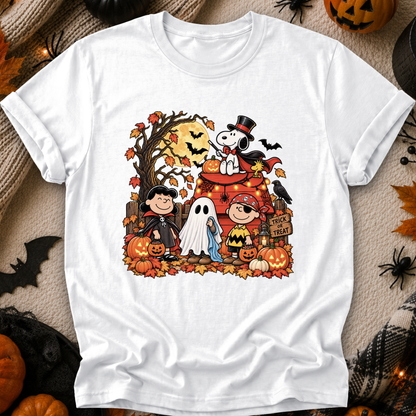 A white short-sleeve crewneck t-shirt featuring a colorful graphic of Peanuts characters in Halloween costumes, styled in a flat-lay with autumn props like pumpkins, spider decorations, and a black spiderweb.