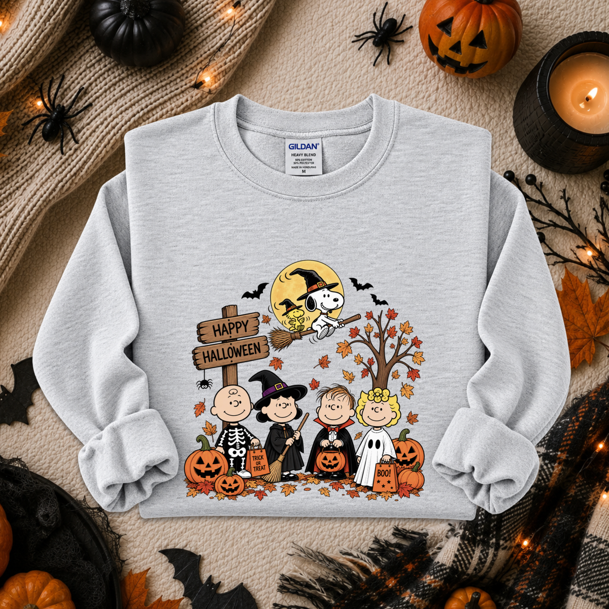 A light heather gray crewneck sweatshirt featuring a colorful 'Happy Halloween' cartoon graphic from The Peanuts Movie, styled in a cozy autumn flat-lay with pumpkins, a candle, and spider decorations.