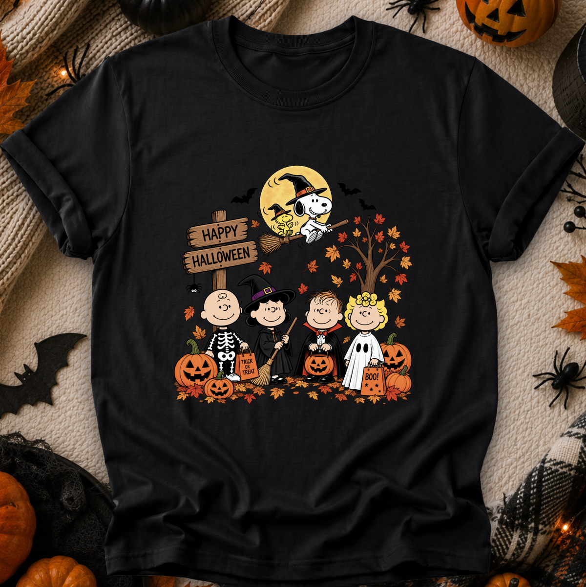 A black short-sleeve graphic T-shirt laid flat on a cozy beige knit blanket with Halloween decorations around it. The shirt features a colorful cartoon print of Peanuts characters dressed in Halloween costumes, pumpkins, a full moon, bats, and a sign reading 'Happy Halloween'.