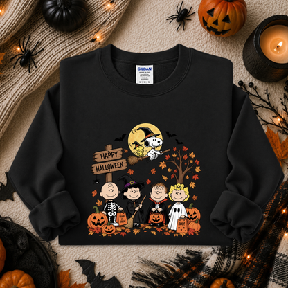 A black crewneck sweatshirt featuring a colorful cartoon graphic of the Peanuts characters in Halloween costumes with pumpkins, a witch on a broomstick, and a 'Happy Halloween' sign. The sweatshirt is displayed in a flat-lay arrangement with autumn props including a candle, a pumpkin, a spider, and a knitted blanket.