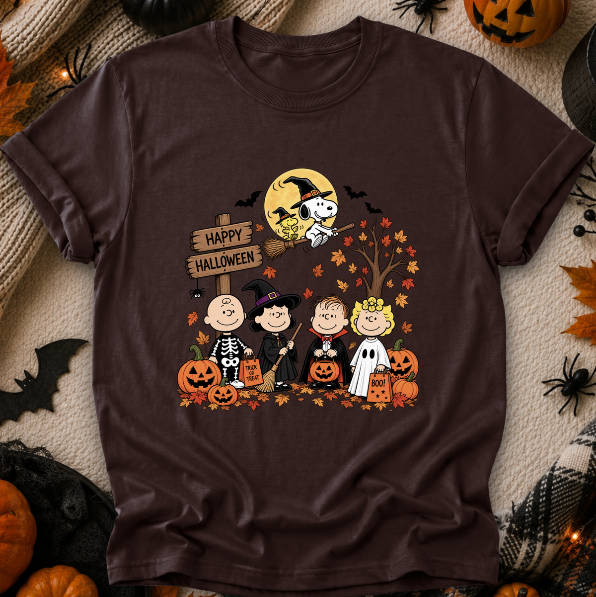 A dark brown short-sleeve t-shirt featuring a colorful 'Happy Halloween' graphic from Peanuts, styled in a flat-lay with seasonal props like pumpkins, a spider, and a plaid blanket.