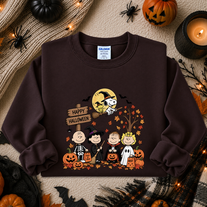 A dark brown crewneck sweatshirt featuring a colorful graphic of the Peanuts characters in Halloween costumes with pumpkins, a full moon, and a 'Happy Halloween' sign. The sweatshirt is displayed in a flat-lay arrangement with seasonal props including pumpkins, a candle, and spider decorations.