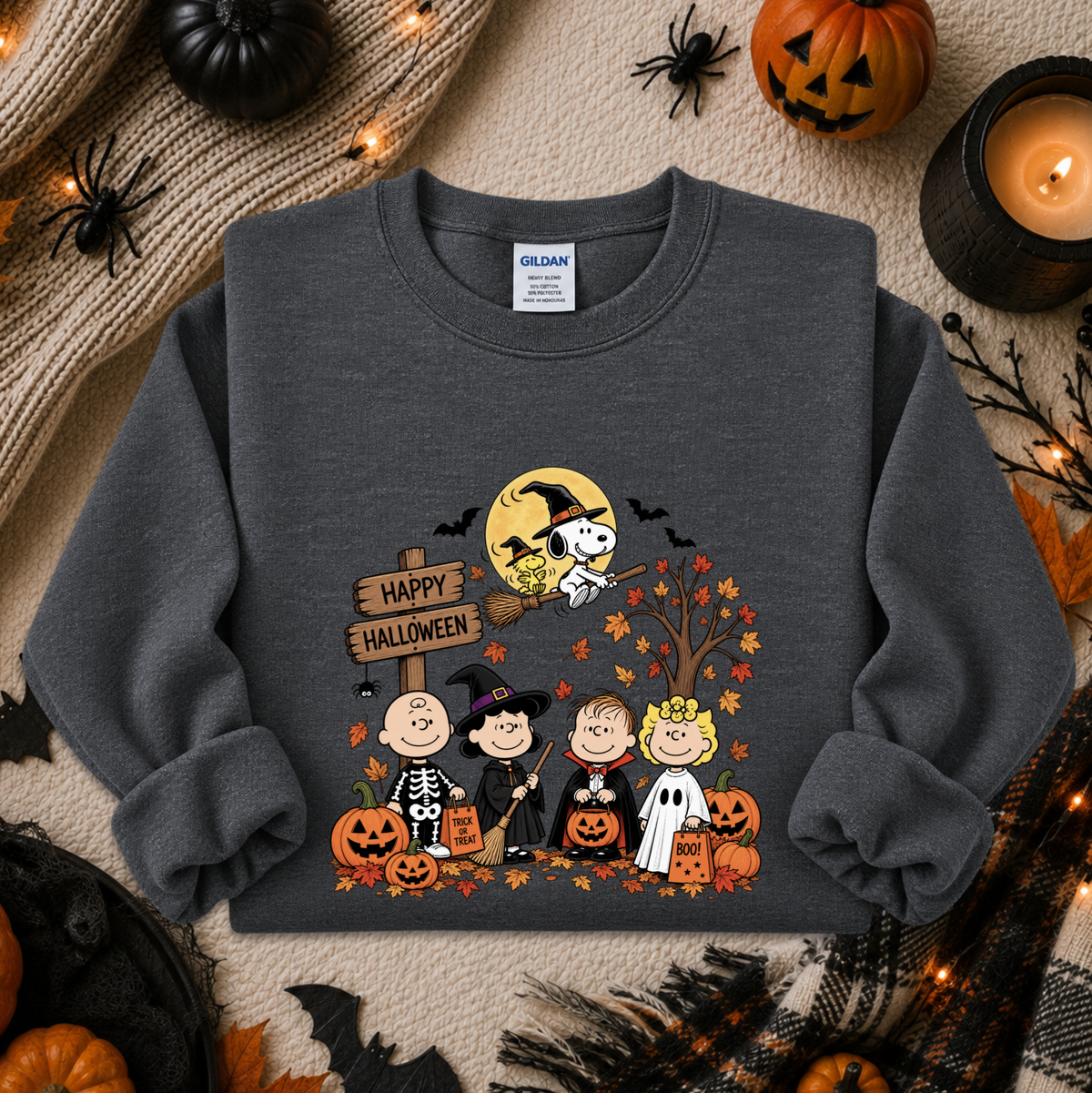 A dark gray crewneck sweatshirt featuring a colorful cartoon graphic of the Peanuts characters in Halloween costumes with pumpkins, a spider, and a 'Happy Halloween' sign. The sweatshirt is displayed in a flat-lay Halloween setting with pumpkins, a candle, and spider decorations.