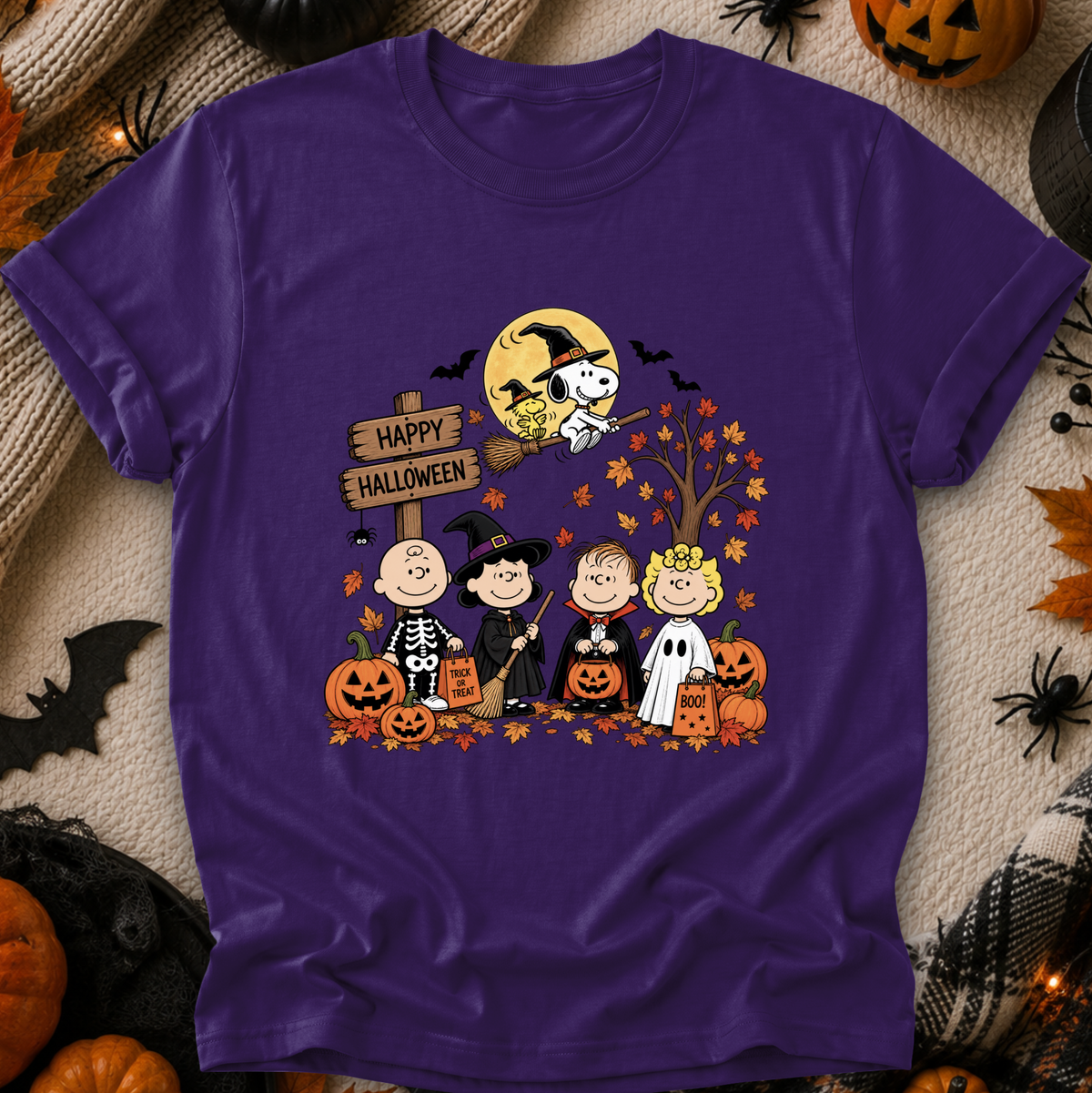 A purple short-sleeve crewneck t-shirt featuring a colorful cartoon graphic of Peanuts characters in Halloween costumes with pumpkins, a full moon, and a 'Happy Halloween' sign, laid flat on a cozy beige knit blanket surrounded by Halloween props.