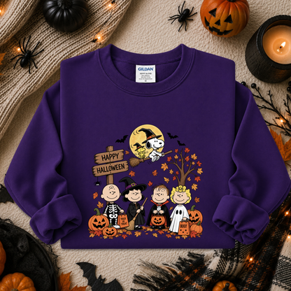 A purple long-sleeve crewneck sweatshirt featuring a colorful 'Happy Halloween' graphic with cartoon characters in costumes, pumpkins, and a witch on a broomstick. The sweatshirt is displayed in a flat-lay Halloween setting with pumpkins, a candle, and spider decorations.