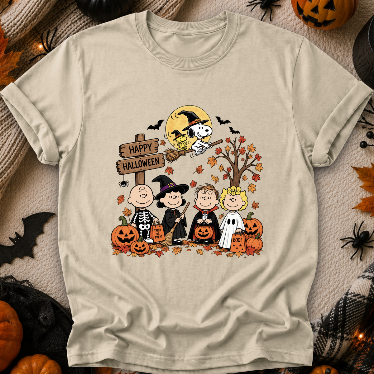 A light beige graphic t-shirt featuring a Peanuts Halloween print with Snoopy on a broomstick and Charlie Brown, Woodstock, Linus, and Sally in costumes, surrounded by pumpkins and autumn leaves. The shirt is displayed flat on a cozy, styled Halloween background with props like plastic spiders, a pumpkin, and a black spiderweb.