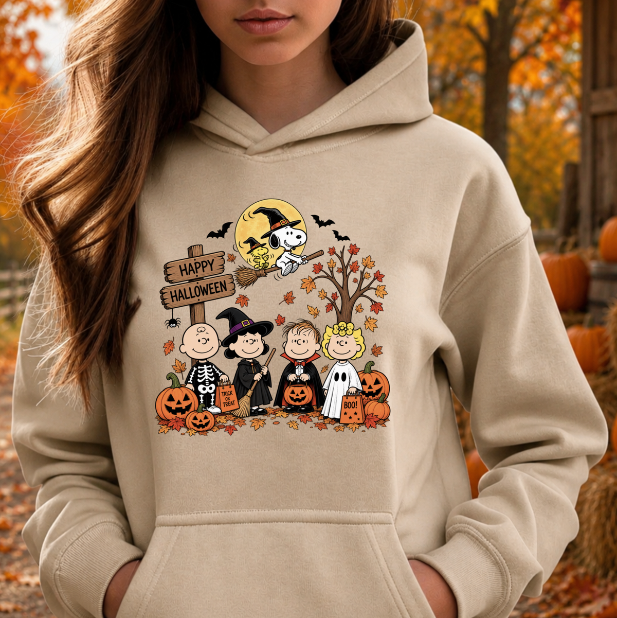 A woman wearing a beige hoodie with a colorful cartoon Halloween graphic featuring Peanuts characters, shown in an outdoor autumn setting with pumpkins and fall leaves.