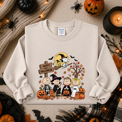 A cream-colored crewneck sweatshirt featuring a colorful 'Happy Halloween' graphic with Peanuts characters, Snoopy, and a doghouse, styled in a cozy autumn flat-lay with pumpkins, a candle, and spider decorations.
