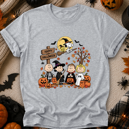 A heather gray short-sleeve crewneck t-shirt featuring a colorful cartoon graphic of Peanuts characters in Halloween costumes, set against a cozy autumn-themed background with pumpkins, spider decorations, and a 'Happy Halloween' sign.