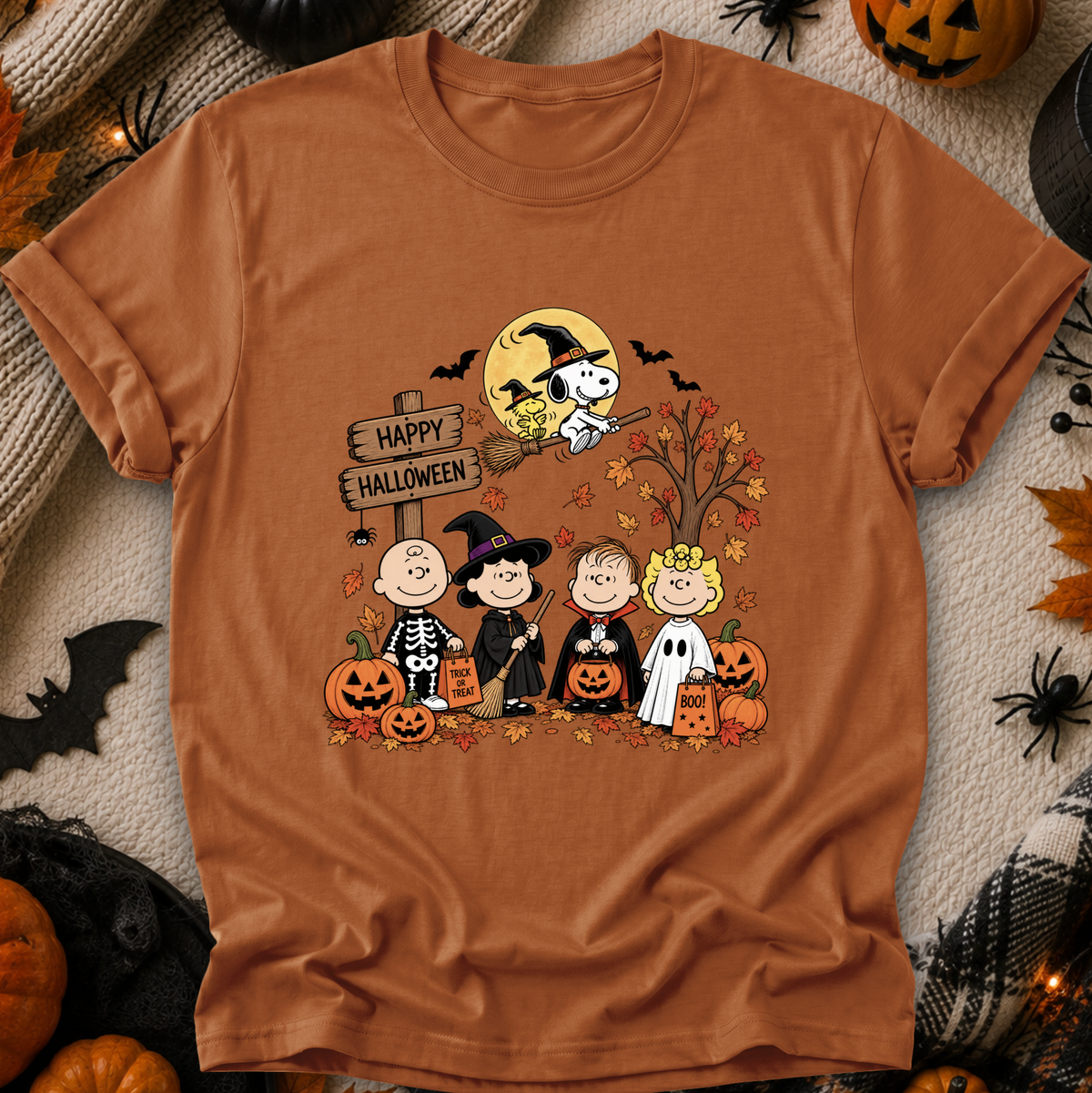 A brown short-sleeve crewneck t-shirt featuring a colorful cartoon graphic of Peanuts characters dressed in Halloween costumes, set against a cozy autumn-themed flat-lay background with pumpkins, spider decorations, and a plaid blanket.