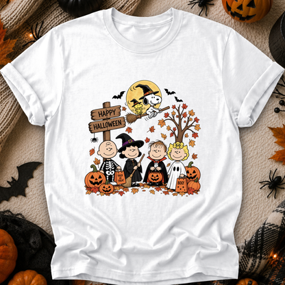 A white short-sleeve crewneck t-shirt featuring a colorful 'Happy Halloween' graphic from Peanuts, styled in a flat-lay with autumn props like pumpkins, spiderwebs, and a spider on a beige textured background.