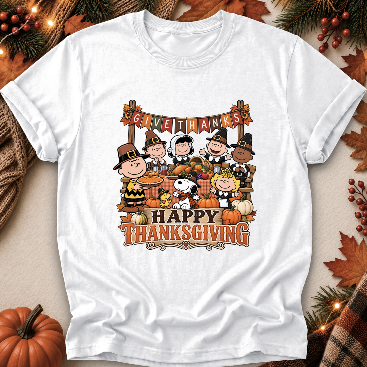 A white short-sleeve graphic T-shirt laid flat on a warm autumn-themed background. The shirt features a colorful Thanksgiving illustration with Peanuts characters, pumpkins, fall leaves, and the text 'GIVE THANKS' and 'HAPPY THANKSGIVING.