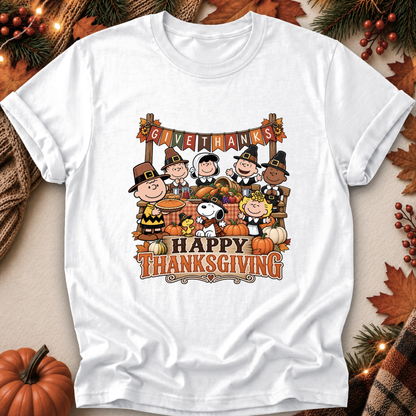A white short-sleeve graphic T-shirt laid flat on a warm autumn-themed background. The shirt features a colorful Thanksgiving illustration with Peanuts characters, pumpkins, fall leaves, and the text 'GIVE THANKS' and 'HAPPY THANKSGIVING.